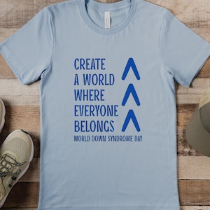 Könnte beinhalten: Hellblaues T-Shirt mit dem Text "Create a world where everyone belongs" und drei blauen Dreiecken. Der Text "World Down Syndrome Day" ist unter den Dreiecken gedruckt.