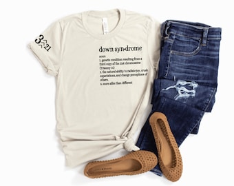 Puede incluir: Una camiseta beige con el texto "down syndrome" definido como una condición genética que resulta de una tercera copia del cromosoma 21 (Trisomía 21). El texto también dice que las personas con síndrome de Down tienen una capacidad natural para irradiar alegría, romper expectativas y cambiar las percepciones de los demás. El texto también dice que las personas con síndrome de Down son más parecidas que diferentes. La camiseta tiene el número 3^21 en la manga.
