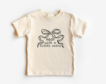 May include: A cream-coloured short-sleeved t-shirt with a black bow and the text "made with a little extra".