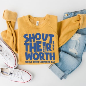 May include: Mustard yellow sweatshirt with the text "SHOUT THEIR WORTH" in blue, along with "WORLD DOWN SYNDROME DAY". The sweatshirt is paired with light blue jeans and white sneakers with red trim.