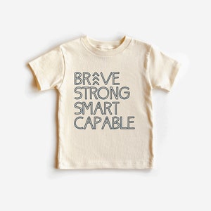 May include: A cream-colored short-sleeved t-shirt with the words "BRAVE STRONG SMART CAPABLE" printed in black on the front.