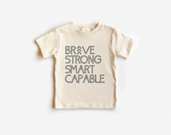 May include: A cream-colored short-sleeved t-shirt with the words "BRAVE STRONG SMART CAPABLE" printed in black on the front.