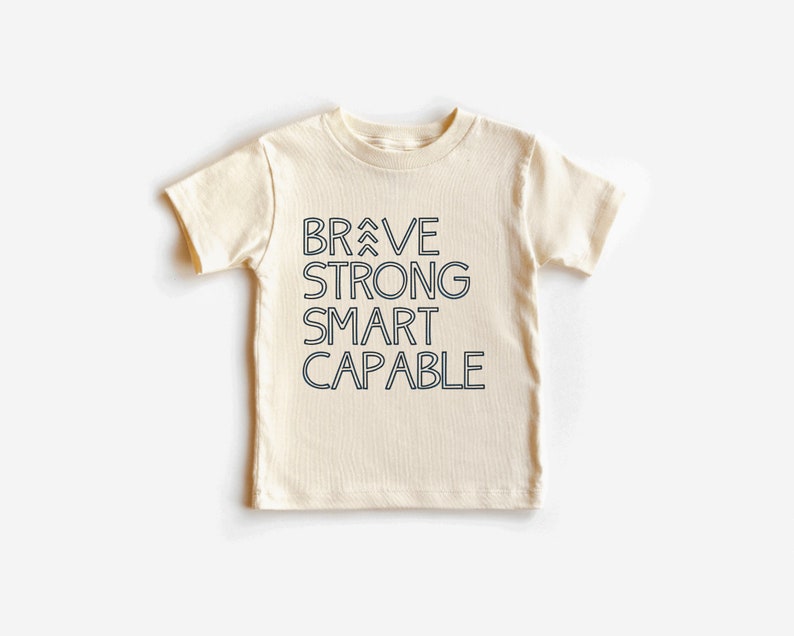 May include: A cream-colored short-sleeved t-shirt with the words "BRAVE STRONG SMART CAPABLE" printed in black on the front.
