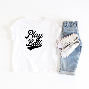 May include: White t-shirt with black text that says "Play Ball" in a retro baseball font. The shirt is laid flat on a white surface with a pair of blue jeans and white sneakers.