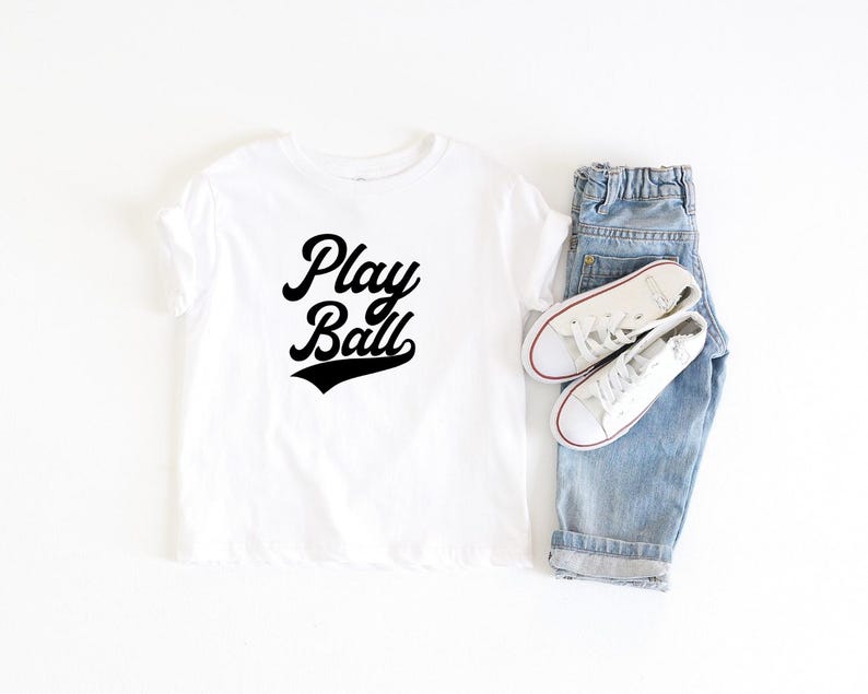 May include: White t-shirt with black text that says "Play Ball" in a retro baseball font. The shirt is laid flat on a white surface with a pair of blue jeans and white sneakers.