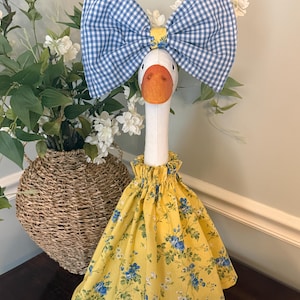 May include: A decorative goose figurine with a white neck and orange beak. It wears a large blue and white gingham bow and a yellow floral dress with blue flowers. The goose is standing on a brown surface.