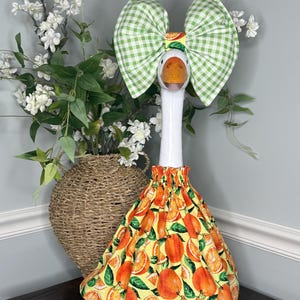 May include: A decorative goose figurine with a white neck and orange beak. It wears a green gingham bow and a skirt patterned with oranges and green leaves on a yellow background. The goose is on a dark wooden surface.