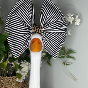 May include: A white goose figurine with a large black and white striped bow. The bow is secured with a leopard print band. The goose has an orange beak and is positioned in front of a basket of flowers.