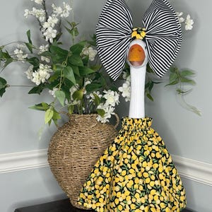 May include: A decorative goose figurine with a large black and white striped bow, a yellow and black lemon-print skirt, and orange feet. A woven basket with white flowers is in the background.