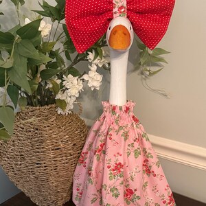 May include: A decorative goose figurine with a white neck and orange beak. It is adorned with a large red polka-dot bow and a pink floral dress. The goose is standing on a small, orange umbrella-like base.