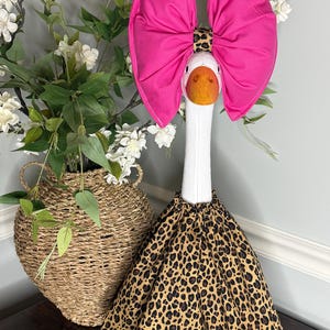 May include: A decorative goose wearing a large pink bow and a leopard print skirt. The goose has a white neck and an orange beak. A wicker basket with white flowers is in the background.