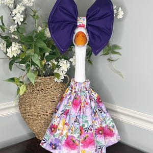 May include: A decorative goose figurine wearing a large purple bow and a floral print dress. The goose has an orange beak and feet. The dress features pink, purple, and yellow flowers on a light purple background. The bow is a deep purple color.