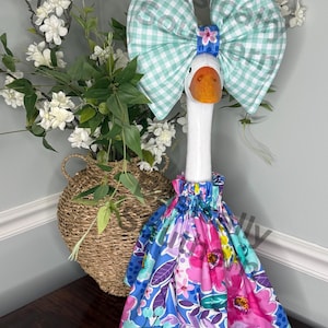May include: A decorative goose figurine with a large, mint green gingham bow and a floral dress. The goose has an orange beak and feet. The dress features a vibrant floral pattern with pink, blue, and purple flowers. The goose is a whimsical home decor item.