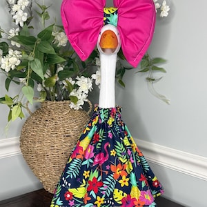 May include: A decorative goose figurine with a large pink bow and a tropical-themed dress. The dress features a dark blue background with colorful floral and flamingo patterns. The goose has a white neck and head with an orange beak and feet.
