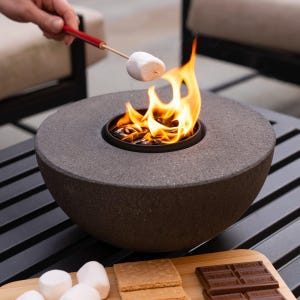 May include: A gray stone fire pit with a small fire burning inside. A marshmallow is being roasted on a stick over the fire. There are graham crackers and chocolate bars on a wooden cutting board next to the fire pit.