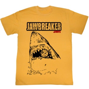 Peut inclure: Un t-shirt jaune avec une illustration en noir et blanc d'une tête de requin avec un cache-œil noir. Le texte "JAWS" est en rouge au-dessus du requin et le texte "JAWSBREAKER" est en noir au-dessus du texte "JAWS".