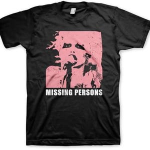 May include: Black t-shirt featuring a pink and black graphic of a person's face. The text "MISSING PERSONS" is printed in white below the graphic. The shirt is made of a soft material.