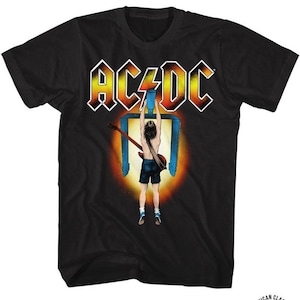 May include: Black t-shirt with a graphic of a man holding a guitar in front of a blue and orange light. The text "AC/DC" is printed in white above the graphic. The text "AMERICAN CLASSICS" is printed in white on a black and white logo in the bottom right corner.
