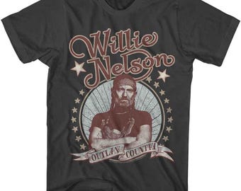 May include: Dark gray t-shirt featuring a portrait of Willie Nelson. The design includes the text "Willie Nelson" in a vintage font, along with "Outlaw Country" on a banner. The image is surrounded by stars.