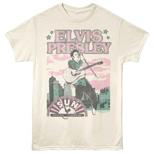 May include: Cream-colored t-shirt featuring a graphic of Elvis Presley playing guitar, with the text "ELVIS PRESLEY" in pink and a cityscape backdrop. The Sun Record Company logo is at the bottom.
