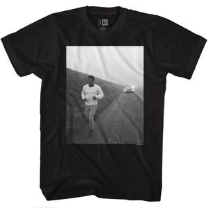 May include: Black t-shirt featuring a photograph of a man running on a foggy road. The photo is in black and white and the man is wearing a white shirt and trousers. The text "ALI" is printed on the shirt.