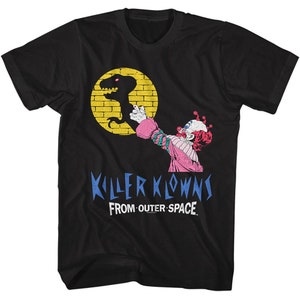 Puede incluir: Camiseta negra con un gráfico de un payaso sosteniendo una silueta de dinosaurio frente a un círculo amarillo con un patrón de ladrillo. El texto "Killer Klowns From Outer Space" está impreso en azul y blanco.