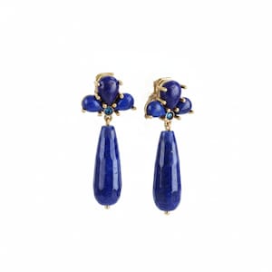 Può includere: Un paio di orecchini pendenti color oro con pietre di lapislazzuli blu intenso. Ogni orecchino presenta un gruppo di tre piccole pietre arrotondate sulla parte superiore con un minuscolo cristallo blu, che sostiene una lunga pietra sfaccettata a goccia.