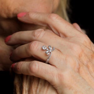 May include: A silver ring with a floral design featuring a large, round, clear gemstone and smaller accent stones. The band is also adorned with small, sparkling stones. The ring is on a person's finger.