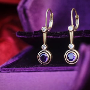 May include: A pair of gold-toned drop earrings featuring a round purple gemstone at the base, a smaller clear stone above it, and a third clear stone near the top lever-back closure. The earrings have a vertical metal bar connecting the stones.