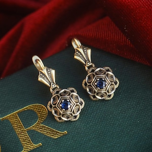 Peut inclure: Une paire de boucles d'oreilles de style ancien. Chaque boucle d'oreille présente un sertissage en métal doré avec une pierre précieuse bleu foncé au centre. Les boucles d'oreilles sont en forme de fleur et sont présentées sur une surface verte avec un fond rouge.