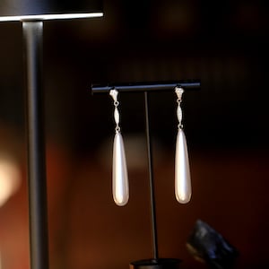 Może przedstawiać: Para białych perełkowych kolczyków wiszących z srebrnym zapięciem i małymi przezroczystymi kamieniami. Kolczyki są prezentowane na czarnym stojaku.