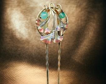May include: A gold-toned hair pin with an Art Nouveau design. The pin features a central circular element with two light green gemstones and stylized leaf-like shapes in pink, white, and gold. Small clear crystals add sparkle.