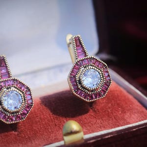 Peut inclure: Une paire de boucles d'oreilles dorées avec un design géométrique. Chaque boucle d'oreille présente une pierre bleu clair centrale entourée d'un anneau de petites pierres rectangulaires de couleur magenta. Les boucles d'oreilles sont présentées dans un écrin en velours rouge.