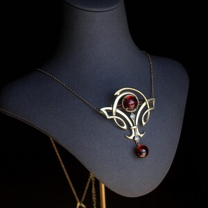 Può includere: Una collana in stile vintage con pendente e catena color oro. Il pendente presenta una grande gemma rossa rotonda al centro, con gemme più piccole e filigrana decorativa. Una gemma rossa più piccola pende sotto.