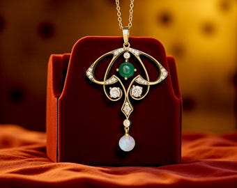 May include: A gold pendant necklace with an Art Nouveau design. The pendant features a central green gemstone, surrounded by clear gemstones and a dangling white pearl. The necklace is displayed on a red velvet box.
