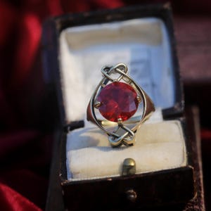 Könnte beinhalten: Ein goldfarbener Ring mit einem großen, runden, facettierten roten Edelstein in der Mitte. Die Ringschiene ist mit komplexen, ineinander verschlungenen Metallschleifen gestaltet, die sich über und unter dem Stein erstrecken und einen dekorativen Rahmen bilden.