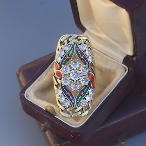 May include: A gold ring with a rectangular shape, featuring a woven design and a central floral pattern. The floral pattern is adorned with clear gemstones, red, green, and blue enamel accents, and is presented in a vintage jewellery box.