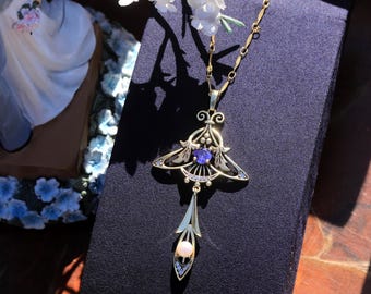 Könnte beinhalten: Eine goldfarbene Anhängerhalskette mit Art-Nouveau-Design. Der Anhänger hat einen zentralen lila Edelstein, kleinere blaue Steine und eine hängende Perle. Die Halskette wird auf einer dunkelblauen Oberfläche präsentiert.