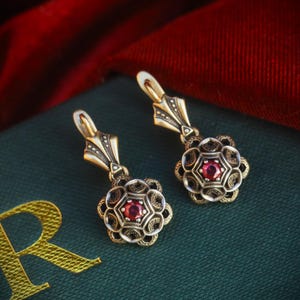 Può includere: Un paio di orecchini in stile antico. Ogni orecchino presenta una gemma rossa centrale circondata da un'incastonatura dettagliata a forma di fiore. Gli orecchini hanno una finitura dorata con dettagli intricati e una chiusura a gancio. Lo sfondo è verde scuro e rosso.