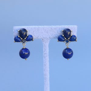 May include: A pair of gold-coloured earrings featuring dark blue gemstones and a hanging round blue stone. The earrings are presented on a white earring holder against a light blue backdrop. The earrings have a classic, elegant design, suitable for various occasions.