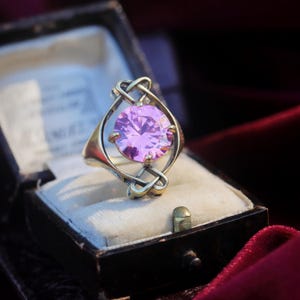 Könnte beinhalten: Ein silberfarbener Ring mit einem runden, facettierten, hellvioletten Edelstein, der in einen dekorativen Metallrahmen mit komplizierten, ineinander verschlungenen, knotenartigen Mustern gefasst ist. Der Ring liegt in einer kleinen Schmuckschatulle.
