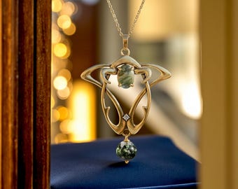 May include: Gold pendant necklace with an Art Nouveau design. The pendant has a central oval green stone, a small diamond, and a round green stone at the bottom. The necklace is displayed on a blue surface.