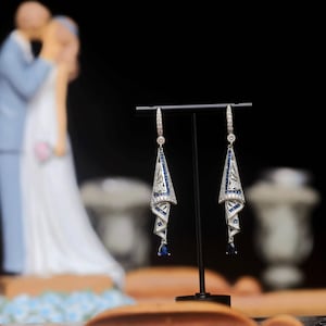Op de afbeelding: Elegante zilveren oorbellen met een geometrisch ontwerp, versierd met witte en blauwe edelstenen. De oorbellen hebben een hangende druppelvorm en worden op een zwarte standaard getoond. Een stijlvol accessoire.