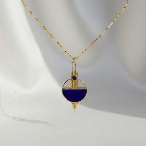 Può includere: Una collana color oro con una catenina sottile e un ciondolo geometrico. Il ciondolo è composto da una barra verticale con una piccola pietra blu circolare in alto e una pietra blu semicircolare più grande alla base, incorniciata da un sottile cerchio dorato.