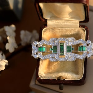 May include: An Art Deco style brooch in silver and green, featuring a geometric design. The brooch is set with clear and emerald green stones.