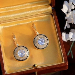 Può includere: Un paio di orecchini pendenti color oro con un cristallo centrale trasparente e un alone di cristalli trasparenti più piccoli. Gli orecchini sono esposti in un portagioie in stile vintage foderato in velluto.
