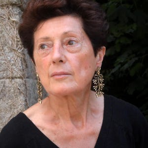 Puede incluir: Una persona mayor con pelo castaño corto y una parte superior negra. La persona lleva pendientes dorados y ornamentados con un diseño en forma de hoja. El fondo incluye una pared de piedra y follaje verde.