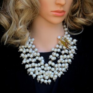Puede incluir: Un collar de perlas llamativo con múltiples hilos de perlas blancas de imitación. El collar está adornado con un broche de insecto dorado con detalles verdes y marrones, agregando un toque único al diseño clásico. El collar se exhibe en un maniquí.