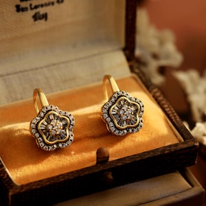 Può includere: Un paio di orecchini a forma di fiore color oro con un gruppo centrale di piccole pietre trasparenti e un alone di pietre più piccole. Gli orecchini sono esposti in un portagioie vintage con interno in velluto.