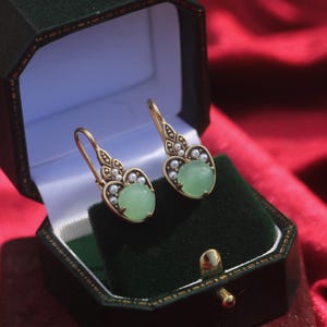 May include: Gold-coloured earrings featuring a pale green gemstone and small white pearls. The earrings are presented in a dark green jewellery box against a red backdrop.
